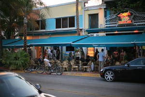 bicycle, building, cafe, canopy, chair, crowd, dusk, eye level view, Florida, Miami, object, people, sign, sitting, standing, street, summer, table, The United States, transport, tree, vegetation, winter